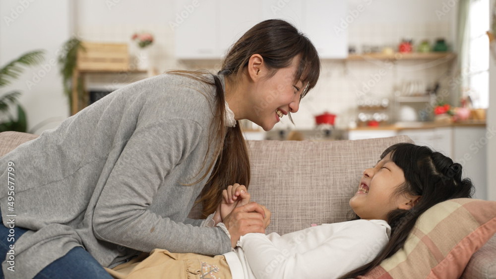 Asian mother and daughter having fun playing tickle together on living room couch at home. the girl pushes her mom away while laughing with excitement