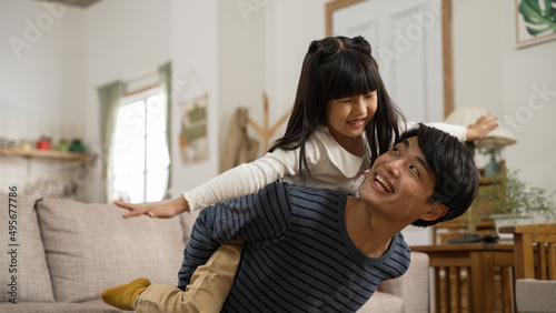 cheerful asian father carrying his cute daughter on back at home. the excited girl is pretending to fly like a plane with open arms
