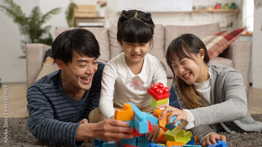 Obraz premium smiling Korean family of three having fun with building block together on ground at home. playful father and mother holding toys attacking each other beside the calm girl