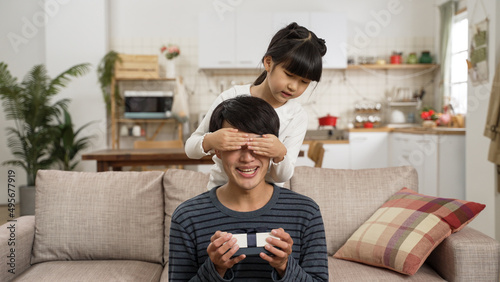asian girl covering father’s eyes and giving him a gift box from behind on father's day at home. she asks her dad to guess what is inside as the man is holding and feeling the present