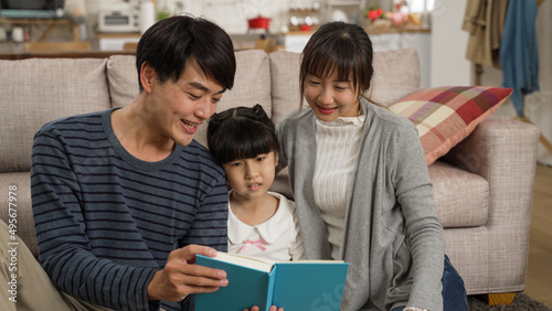 cheerful asian family of three enjoying reading together in the living room at home. the smiling father is having fun telling story with a book in hand.
