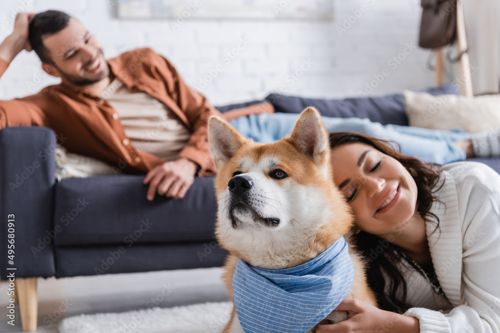 smiling young woman hugging akita inu dog near blurred boyfriend on couch.