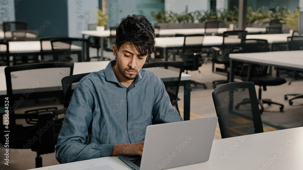 Young man Arabian employee writer thoughtfully typing laptop keyboard copying text in electronic form focused man checking original documents writing new article for website checking figures in report Young man Arabian employee writer thoughtfully typing laptop keyboard copying text in electronic form focused man checking original documents writing new article for website checking figures in report