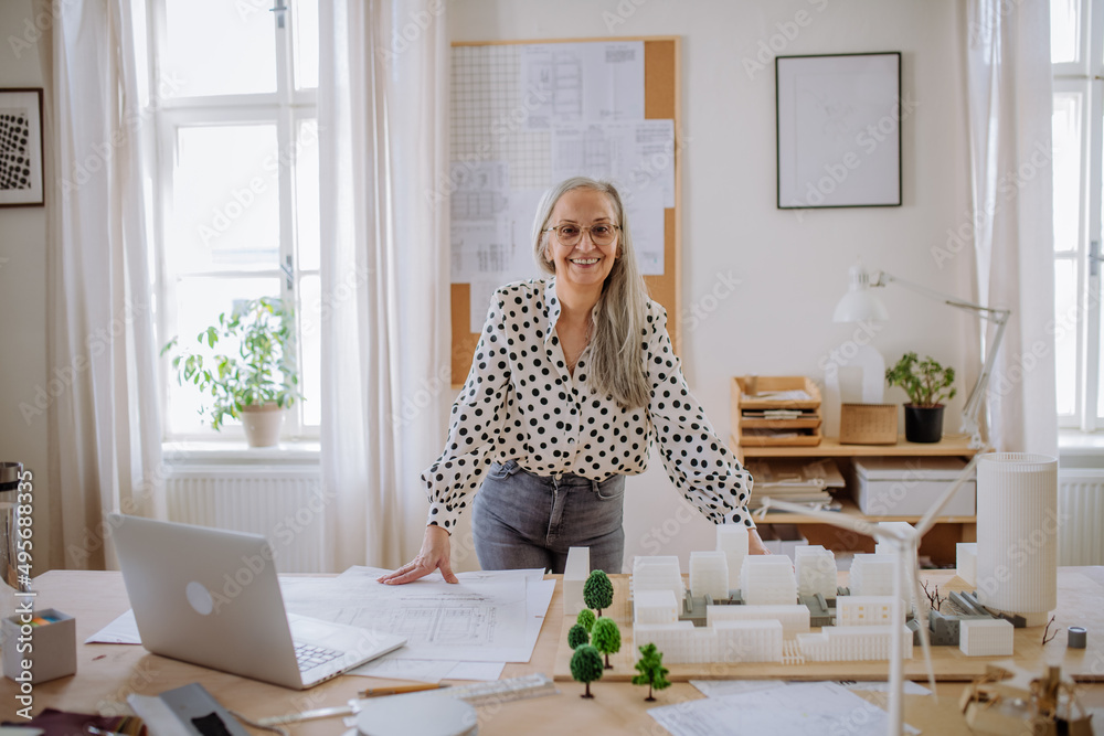 Mature woman architect with model of houses standing in office and looking at camera. Mature woman architect with model of houses standing in office and looking at camera.
