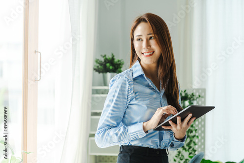 Asian beauty female sit next to window with light glow freelance work at home. Attractive asian woman happiness working with tablet in business casual dress.