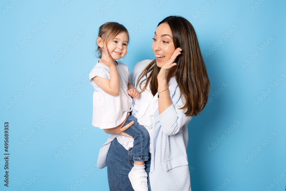 Photo of pretty young mommy hugging little daughter standing isolated over blue background. Motherhood concept. Photo of pretty young mommy hugging little daughter standing isolated over blue background. Motherhood concept.