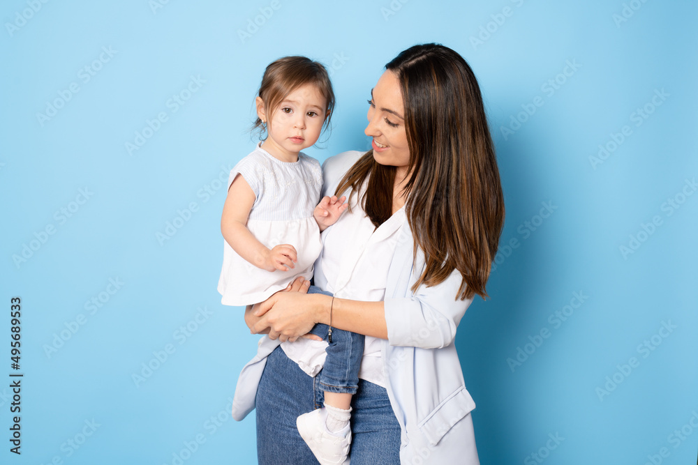 Photo of pretty young mommy hugging little daughter standing isolated over blue background. Motherhood concept.