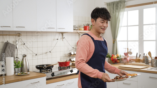 cheerful asian househusband wearing apron is preparing meal for family in a bright kitchen at home. he put several healthy dishes on dining table