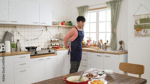 happy asian dad just finishing preparing lunch for family in a modern bright kitchen at home. he puts the last dish on the table and removes his apron