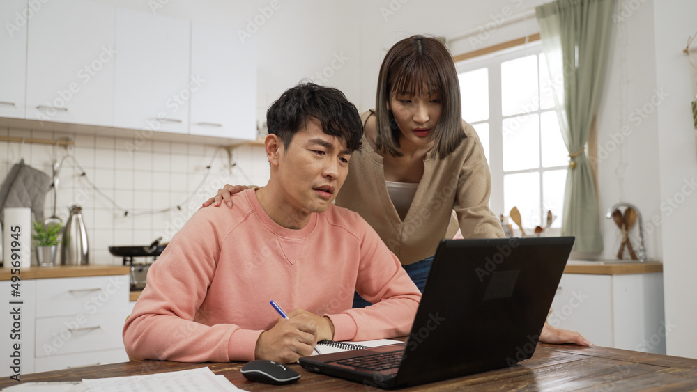 unhappy Asian man feeling anxiety while working from home on computer in the dining room. his wife walking near to comfort him while he is taking notes unhappy Asian man feeling anxiety while working from home on computer in the dining room. his wife walking near to comfort him while he is taking notes