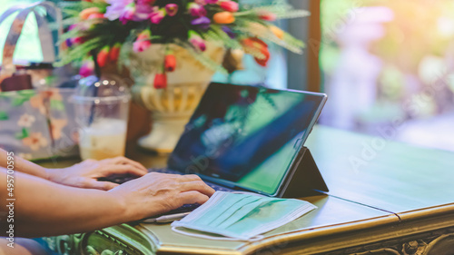 Fotografie Hands of mature woman typing on laptop keyboard to work from home and take off her surgical mask put it on the table for easy breathing