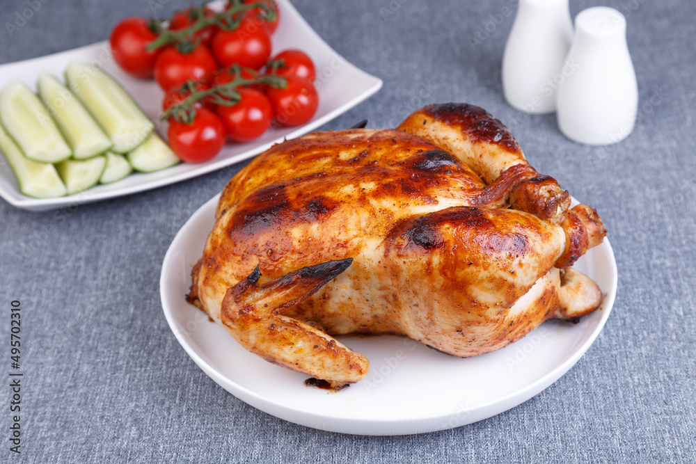 Chicken baked whole to a delicious crust. A traditional dish. In the background are cucumbers and cherry tomatoes on a white plate. Close-up, selective focus, gray background. Chicken baked whole to a delicious crust. A traditional dish. In the background are cucumbers and cherry tomatoes on a white plate. Close-up, selective focus, gray background.