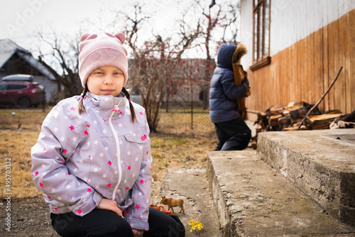 Little girl in warm clothes sitting close to simple wooden house in village in spring outdoors