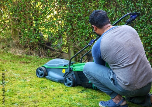 An unrecognizable asian indian man sitting on grass and adjusting the grass basket from a electric lawn mower machine.