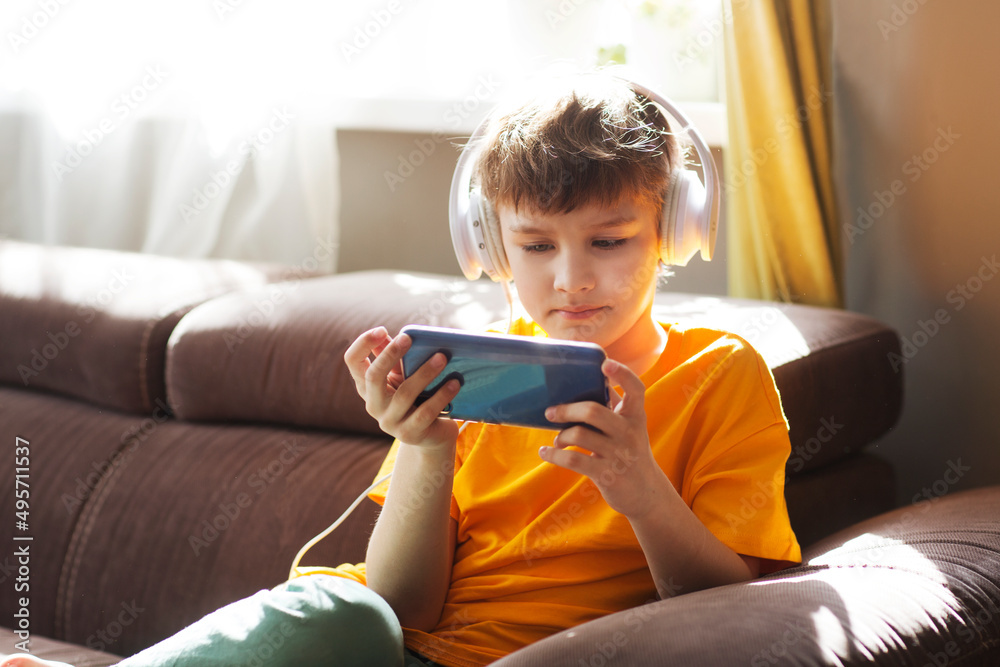 kid use the phone, play the game. teenager in headphones plays on a smartphone or watches video sitting on the couch at home kid use the phone, play the game. teenager in headphones plays on a smartphone or watches video sitting on the couch at home