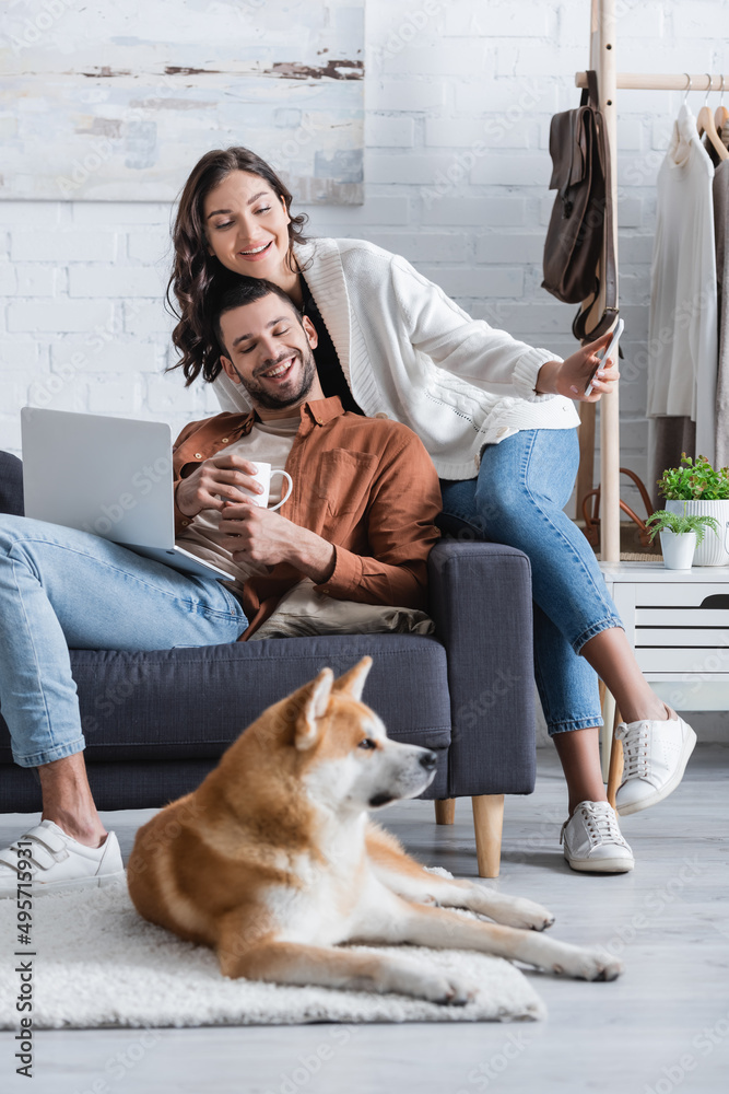 smiling young woman holding smartphone near boyfriend with laptop and looking at akita inu dog in living room.