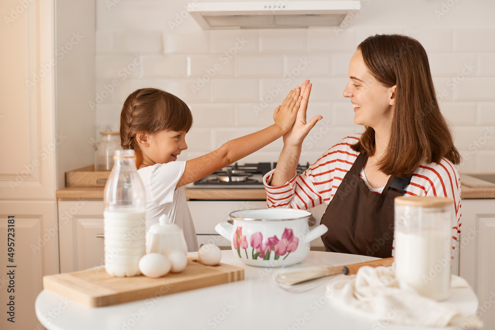 Indoor shot of mother and her cute charming little daughter playing and giving five to each other while baking in kitchen at home, surrounded with bake ingredients. Indoor shot of mother and her cute charming little daughter playing and giving five to each other while baking in kitchen at home, surrounded with bake ingredients.