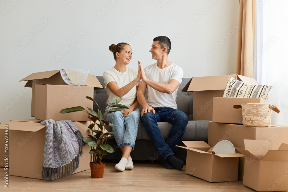 Portrait of attractive young couple sitting on sofa surrounded with carton boxes, family during relocation into a new flat, holding hands, looking at each other, expressing happiness. Portrait of attractive young couple sitting on sofa surrounded with carton boxes, family during relocation into a new flat, holding hands, looking at each other, expressing happiness.