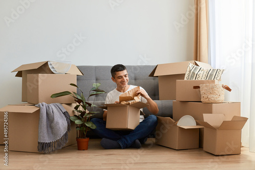 Horizontal shot of brunette positive man wearing white t shirt sitting on floor near sofa surrounded with cardboard boxes, upacking his personal piles after moving in. photo