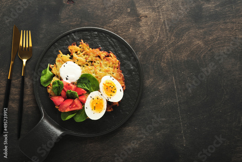 Potatoes latkes with sour cream, spinach salad, tomatoes and boiled eggs on dark wooden old table background. Homemade tasty potato pancakes. Delicious food for breakfast. Top view.