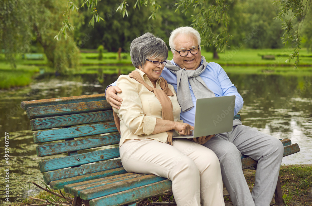 Fototapeta premium Happy elderly retired husband and wife spend time together, sitting on a bench in the park and looking at the laptop screen, communicating with loved ones at a distance. Outdoor recreation.