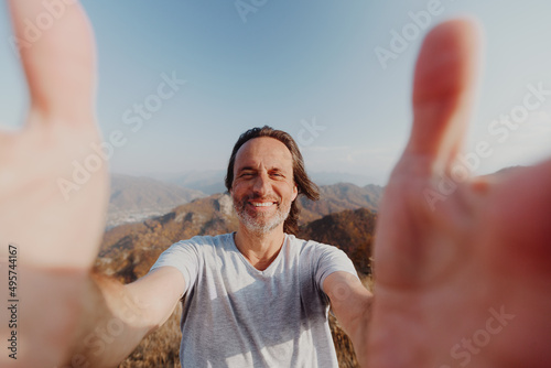 A middle-aged man on top of a mountain takes a selfie. He holds the camera with both hands. He is happy and smiles widely.