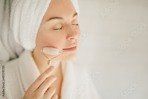 Beautiful young woman with perfect skin wearing white bathrobe and towel on head after shower, making face massage using a jade roller in bathroom.