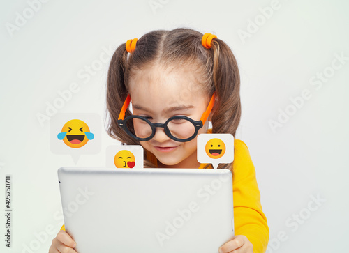 Beautiful cute little girl with glasses using tablet computer on white background. Social media concept.  Child sending kiss and laugh smiley emojis to a friend.