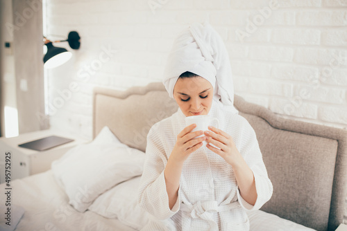 Young woman wearing white bathrobe sitting on bed, drinking hot coffee in her bedroom.