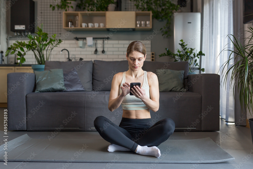 Obraz premium Woman doing yoga at home sitting in lotus position on the floor, using phone for online fitness and yoga classes