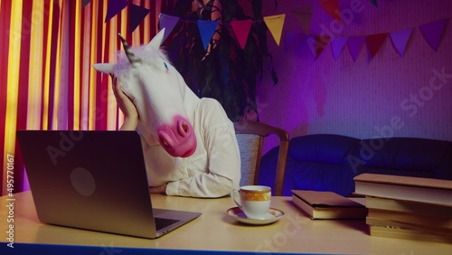 A woman in a unicorn mask is sitting on a chair at a table and typing on a laptop keyboard. This man is desperate. Multicolored lighting. Masquerade