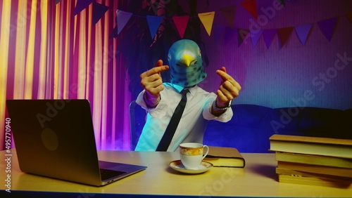 A young man in a pigeon mask sits on a chair at the table and shows a heart sign with his fingers. Multicolored lighting. Masquerade