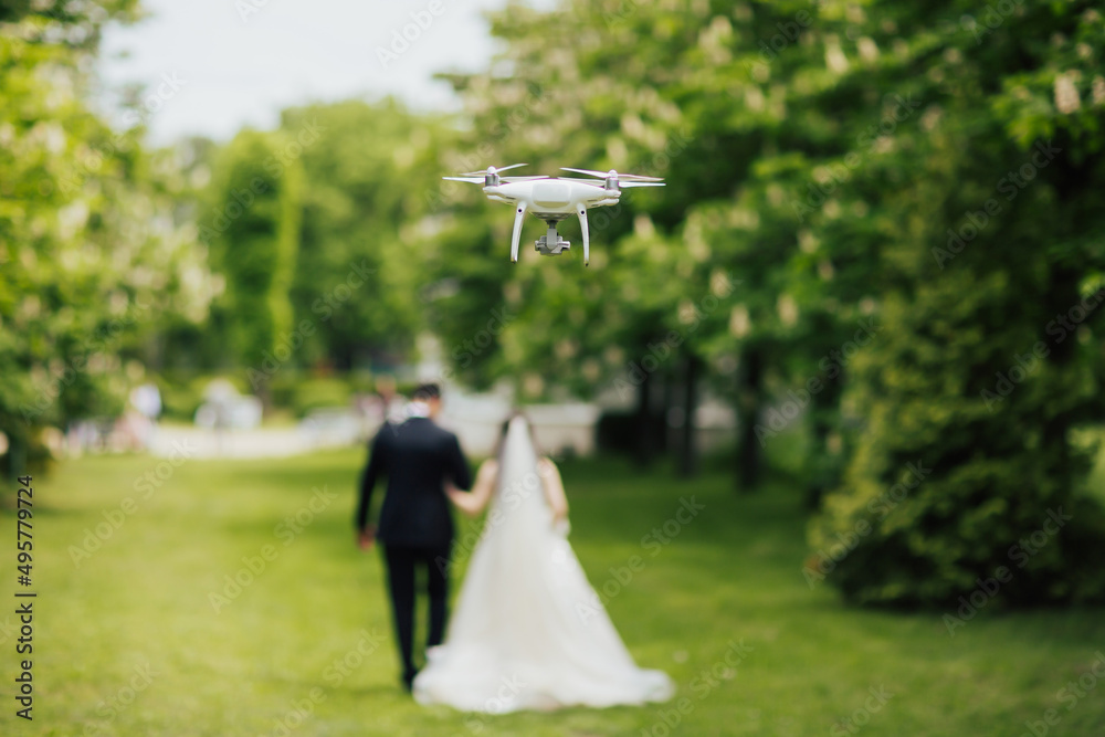 Photo of a drone filming wedding couple while they walk along in the park at wedding day.