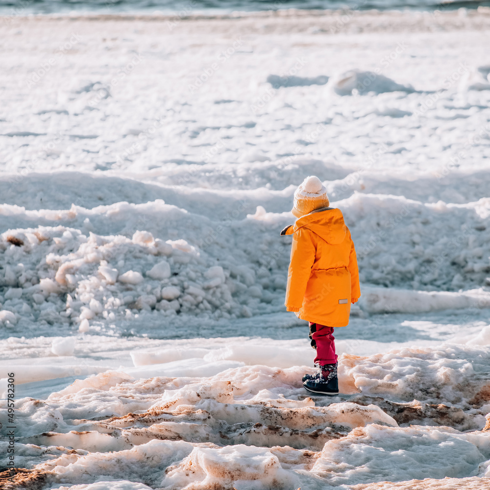 Little girl in yellow jacket walks on snow at sunny winter day.