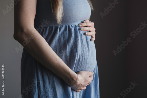 Pregnant woman in dress holds hands on belly. Pregnancy, maternity, preparation and expectation concept. Close-up, copy space, indoors. Beautiful tender mood photo of pregnancy