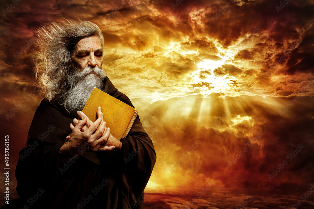 Prophet with Bible. Old Monk with Golden Book praying over Epic Landscape Background. Senior Bearded Man Worship in Black Cloak over Mystery Sunset Sky