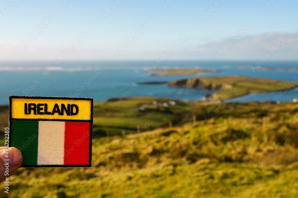 Tourist holding badge with sign Ireland and Irish National flag in focus. Stunning nature scenery out of focus in the background. Nobody. Sky road. county Galway, Ireland.
