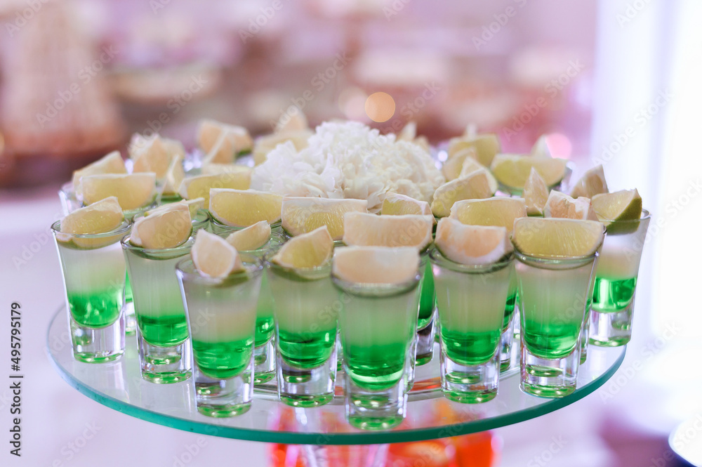 Close view of assorted shots with cocktail, which includes of green beverage, white tequila and piece of lime, located on glass plate against blur background Close view of assorted shots with cocktail, which includes of green beverage, white tequila and piece of lime, located on glass plate against blur background