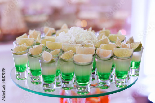 Close view of assorted shots with cocktail, which includes of green beverage, white tequila and piece of lime, located on glass plate against blur background
