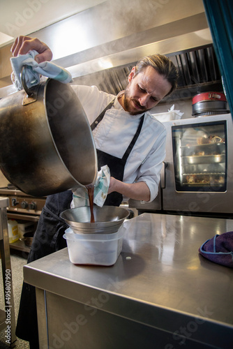 Título: chef rubio con barba en interior de cocina de restaurante filtra salsa de una olla grande metalica con uniforme blanco y mandil negro, manos y close-up photo