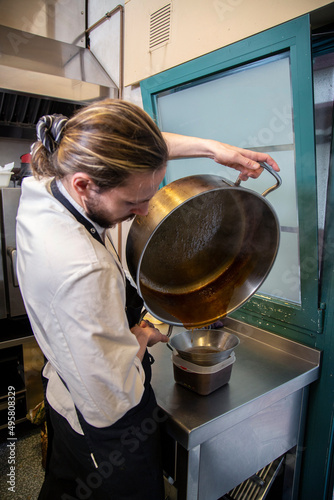 chef rubio con barba en interior de cocina de restaurante filtra salsa de una olla grande metalica con uniforme blanco y mandil negro, manos y close-up photo