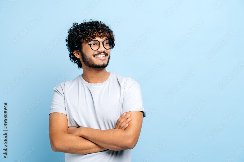 Attractive positive Indian or Arabic curly haired guy with glasses, wearing a basic t-shirt, student or freelancer, standing over isolated blue background, with arms crossed, looks to the side, smiles Attractive positive Indian or Arabic curly haired guy with glasses, wearing a basic t-shirt, student or freelancer, standing over isolated blue background, with arms crossed, looks to the side, smiles