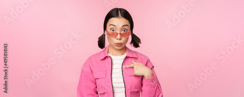 Close up portrait of asian girl looks surprised, points at herself with disbelief, being chosen, stands over pink background