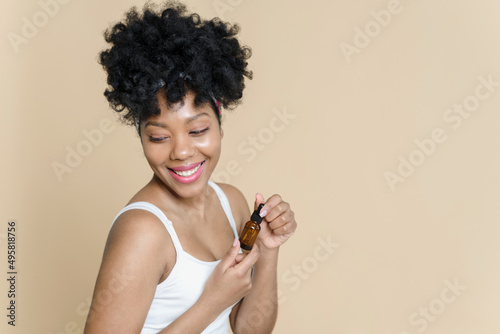 Portrait of a beautiful black woman holding a bottle of moisturizing serum on a beige background. African American young woman smiling while holding a jar of oil.