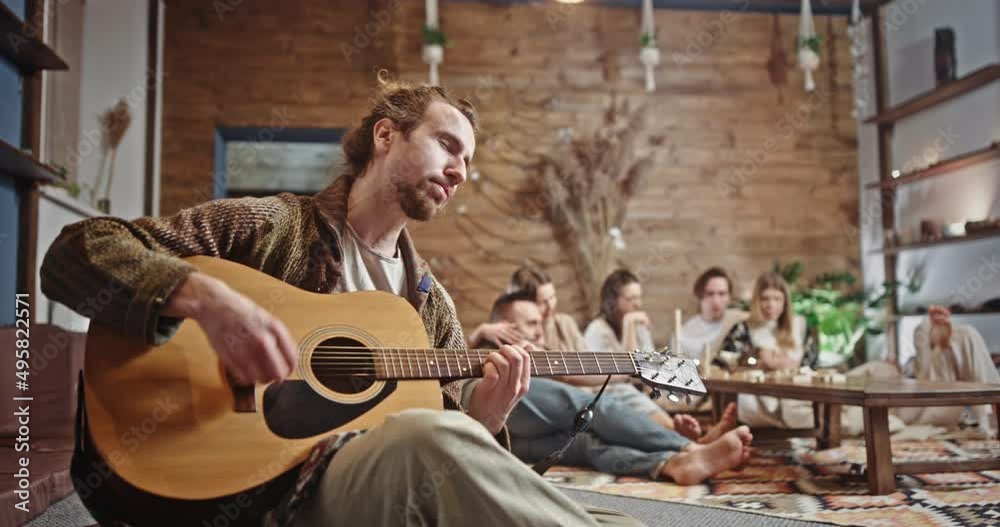 Male hippie playing guitar on home. Low angle of young bearded man playing acoustic guitar during meeting with friends in loft room in weekend