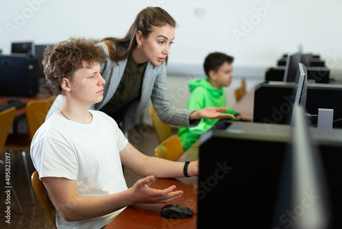 Young female teacher helping focused teenage student studying for exam in college library computer lab..