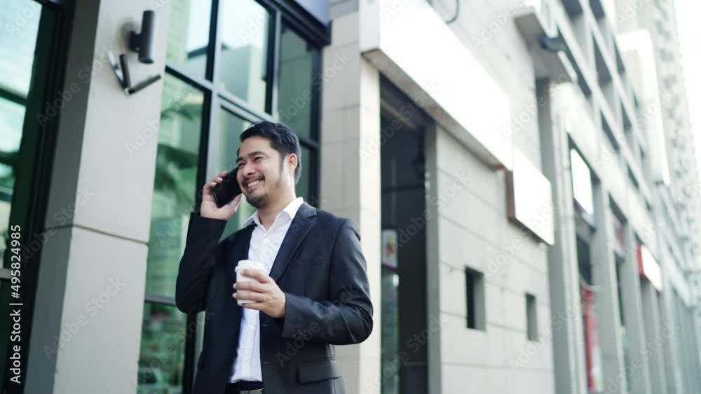 Asian businessman Smiling Wearing a Black Suit is Walking On the Streets Of the City to Work. in Hand holding a Coffee Cup using Smartphone. Urban life Style Concept. Asian businessman Smiling Wearing a Black Suit is Walking On the Streets Of the City to Work. in Hand holding a Coffee Cup using Smartphone. Urban life Style Concept.