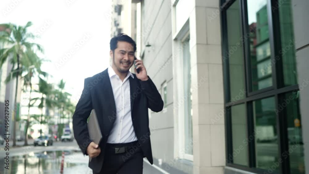 Asian Businessman Smiling Wearing a Black Suit is Rush Running On the Streets Of the City to Work. in Hand holding a Laptop using Smartphone. Urban lifeStyle Concept. Asian Businessman Smiling Wearing a Black Suit is Rush Running On the Streets Of the City to Work. in Hand holding a Laptop using Smartphone. Urban lifeStyle Concept.