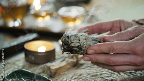 Woman hands burning white sage, before ritual on the table with candles and green plants. Smoke of smudging treats pain and stress, clear negative energy and meditation