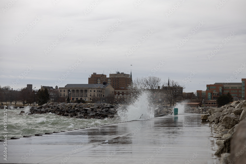 Stormy water splashing up and over the pier. Riprap along the shoreline. Buildings in background with gray cloudy sky. Wind blowing misty water over the rocks and pier. Bare trees along the shoreline.